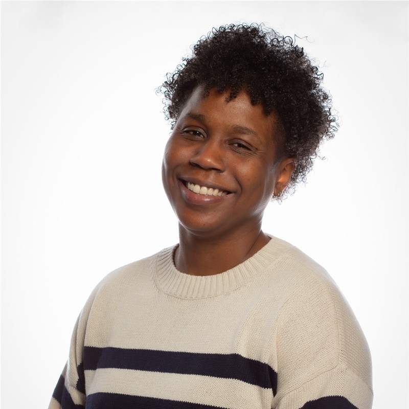 A person with short curly hair smiles at the camera, wearing a cream-colored sweater with dark horizontal stripes, against a plain white background.