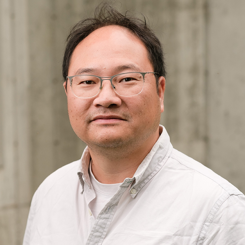 A man wearing glasses and a light-colored button-up shirt stands in front of a plain, light gray concrete wall.