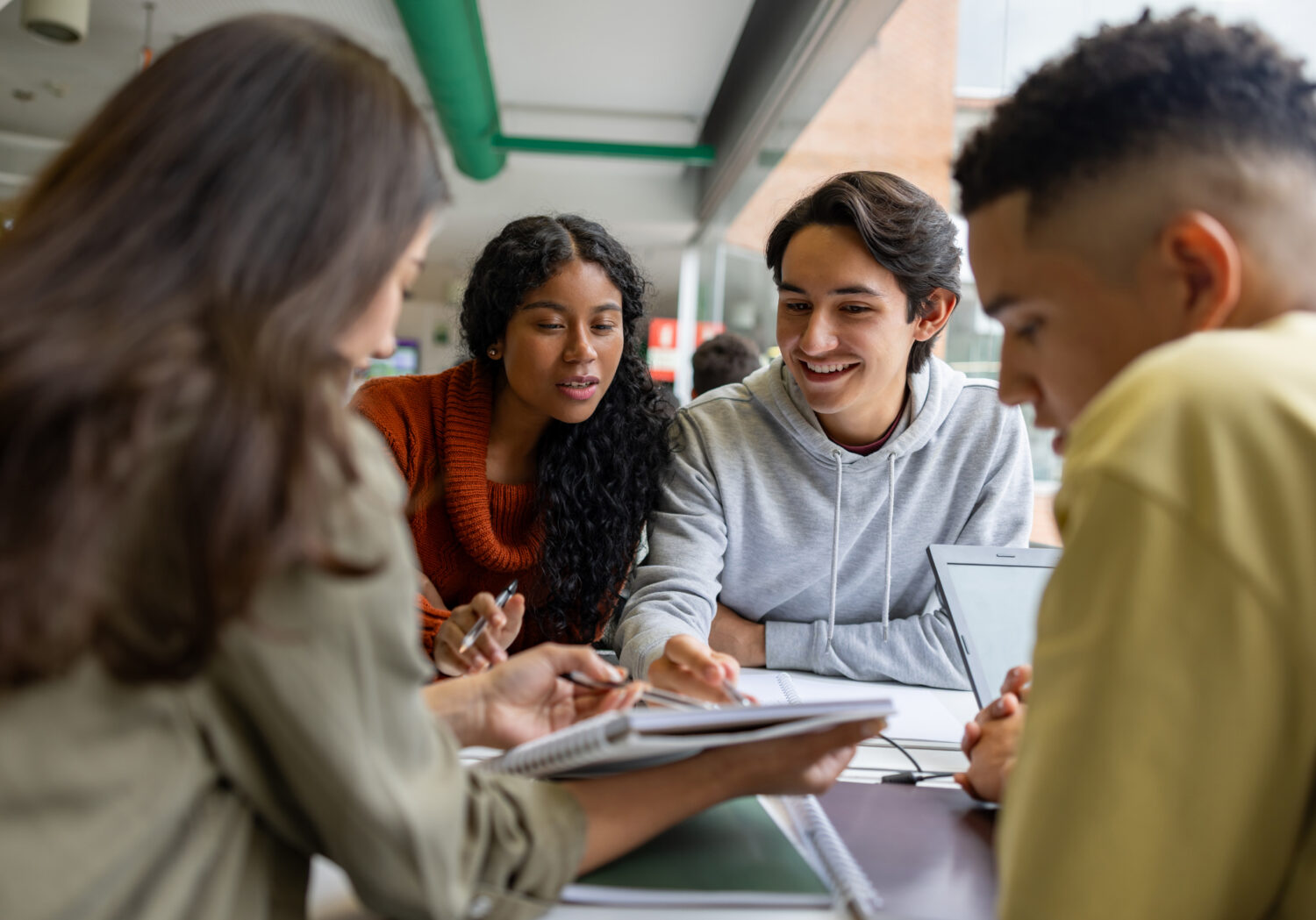 Four young adults sit around a table indoors, looking at documents or notebooks and discussing together. A laptop is also visible on the table.